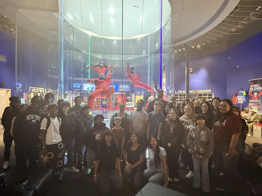 Rudd Middle School STEM students stand together for a photo. They pose at an indoor skydiving facility. Behind them, instructors soar through the air while inside the wind tunnel.