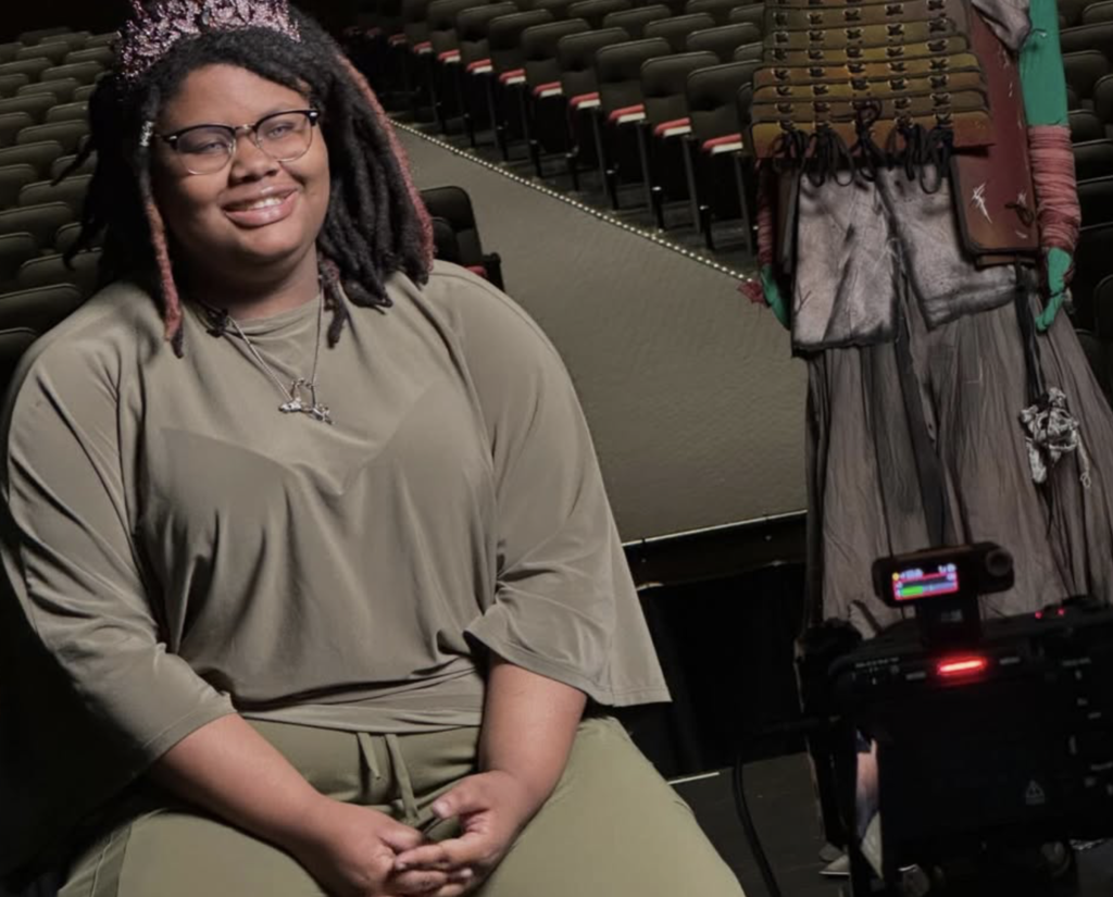 A student smiles while doing an interview in front of a camera. The student sits on a stage in a school auditorium.