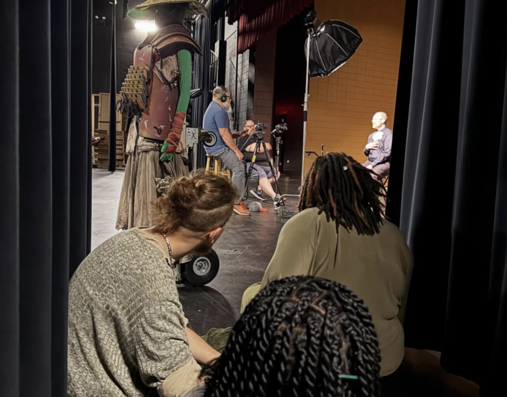 Students watch from the curtains as a man does a news interview on stage in a school auditorium.