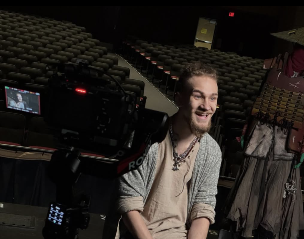 A student sits on stage in an auditorium and answers questions while doing an interview with the news.