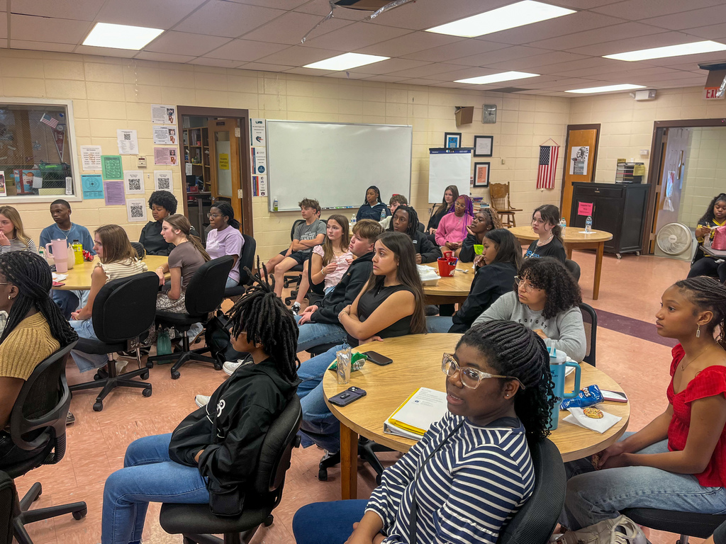 Students sit at tables in a room and look forward while listening to someone speak during the Superintendent's Student Advisory Council meeting.