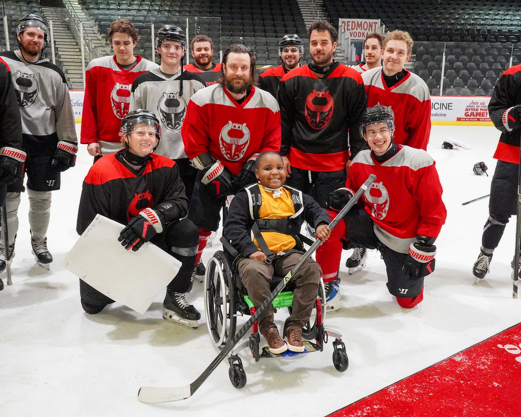A student in a wheelchair holds a hockey stick. He is surrounded by members of the Birmingham Bulls hockey team.