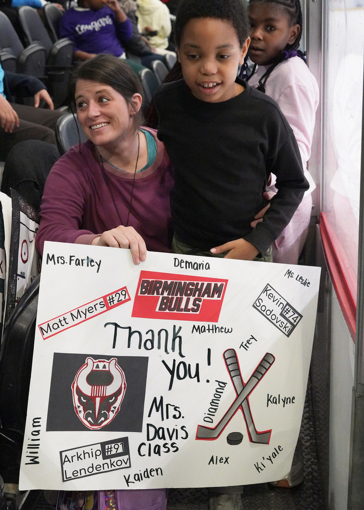 A student and a teacher are sitting in the stands during a Birmingham Bulls hockey practice. They are smiling and holding up a sign celebrating the Bulls players.