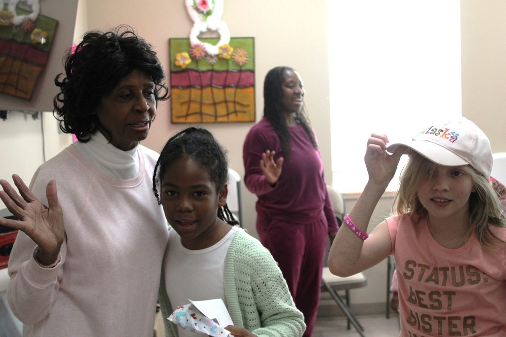 A group of girls and women stand in a room. One of the women hugs a little girl.