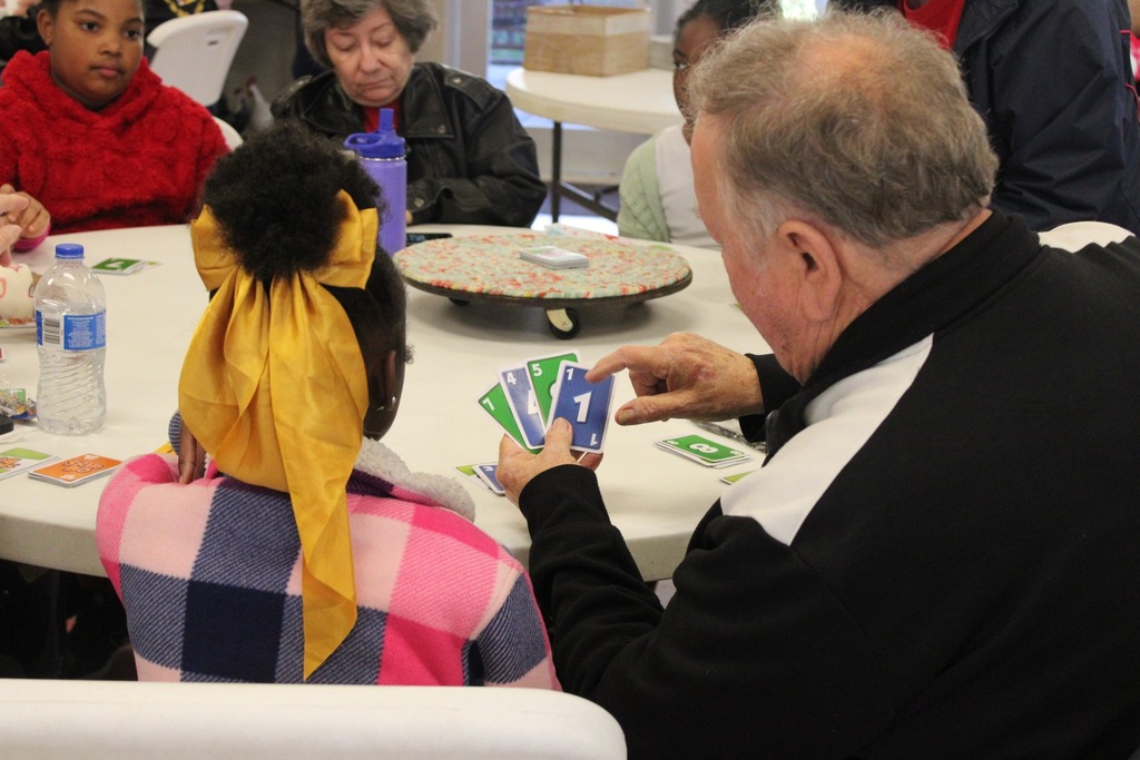 A group of children and adults sit at a table. One of the men at the table shows a little girl a stack of cards.