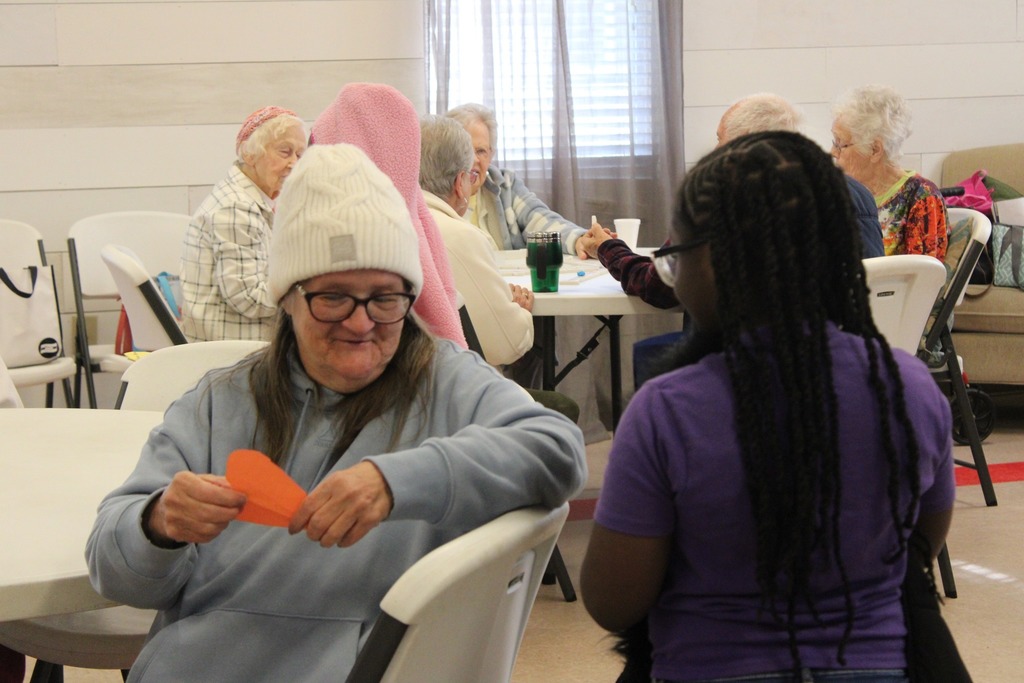 A girl talks to a woman inside a senior center. The woman holds a homemade card.