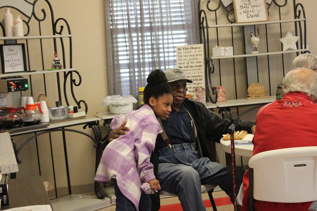 A girl hugs a man while they sit and stand inside a senior center.