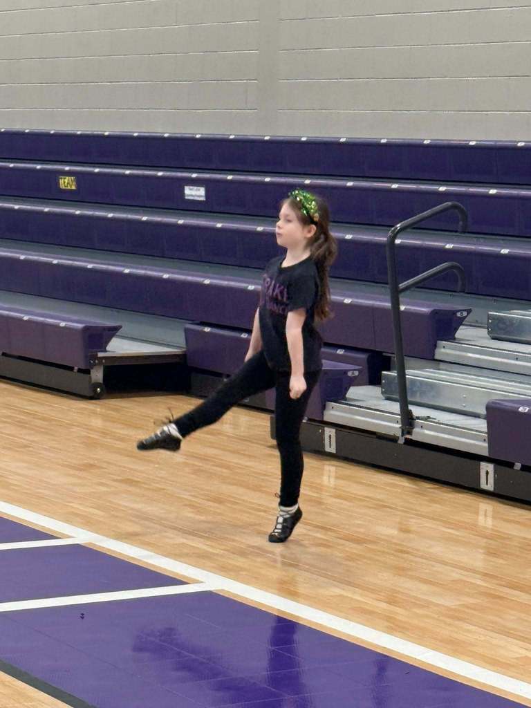 A girl performs an Irish dance inside a school gym. The girl wears an all black outfit with a  sparkly green headband and dance shoes.