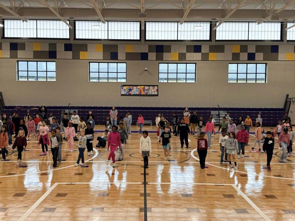 A large group of students stand in a gym and learn a dance.