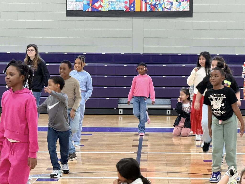 Students stand in lines in a school gym and learn a dance routine.