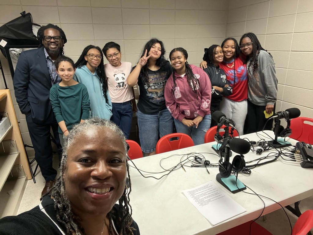 Two adults and a group of students stand together and take a selfie. They stand in a room that has a table in the middle with podcast equipment on it.