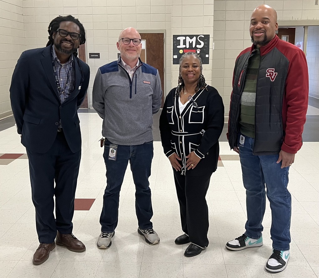 Four adults stand together for a photo and smile. They stand in the Irondale Middle School hallway.
