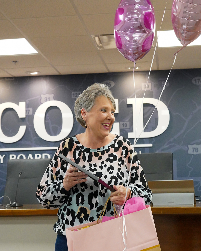 A woman laughs while holding up pink balloons, a pink gift bag, and a certificate. 