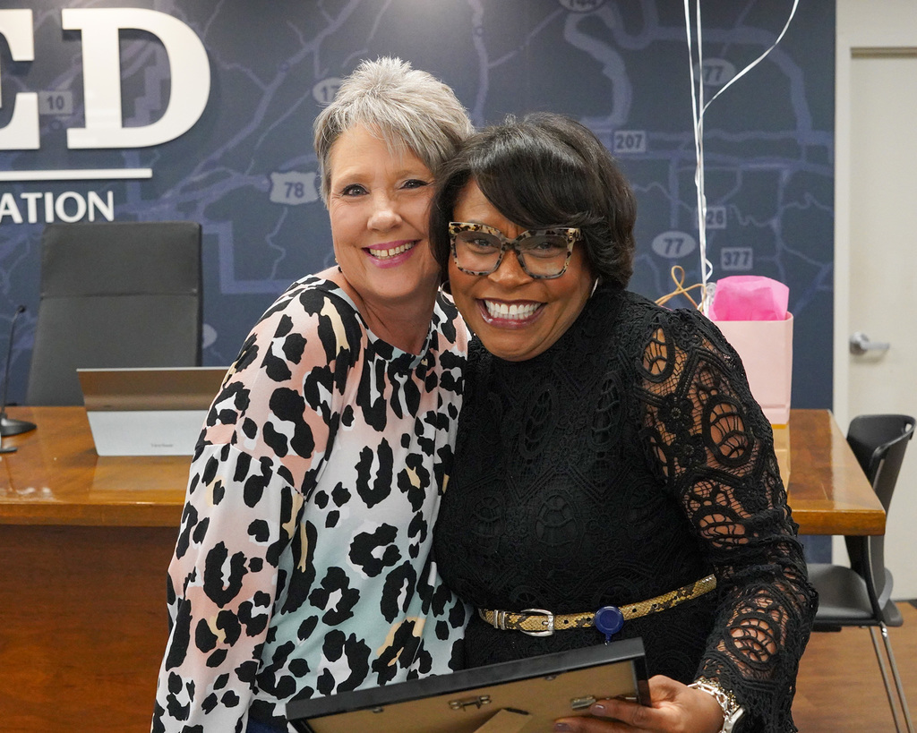 Two women give each other a side hug and smile widely at the camera. The woman on the left side of the image is being presented an award by the woman on the right side of the image. 