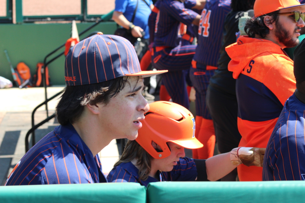 A player from the Fultondale High School baseball team looks onto the field during the 4th Annual Rickwood Classic at Historic Rickwood Field in Birmingham, AL. He appears to be hot and is wearing his baseball cap. Other players are behind him. 