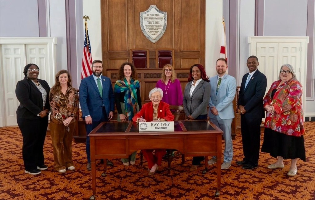 Teachers and educational leaders stand together for a photo inside a room. They stand behind Alabama Governor Kay Ivey as she sits at a table and holds a pen.