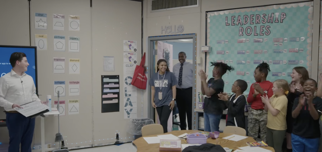 Clay Elementary School teacher Ms. Joy Isom walks into a classroom and smiles. Principal Dr.  Jacqnaii Finkley walks in behind her and smiles too. Students stand in the classroom and clap for Ms. Isom. A man also stands in the classroom and smiles while holding a big check.