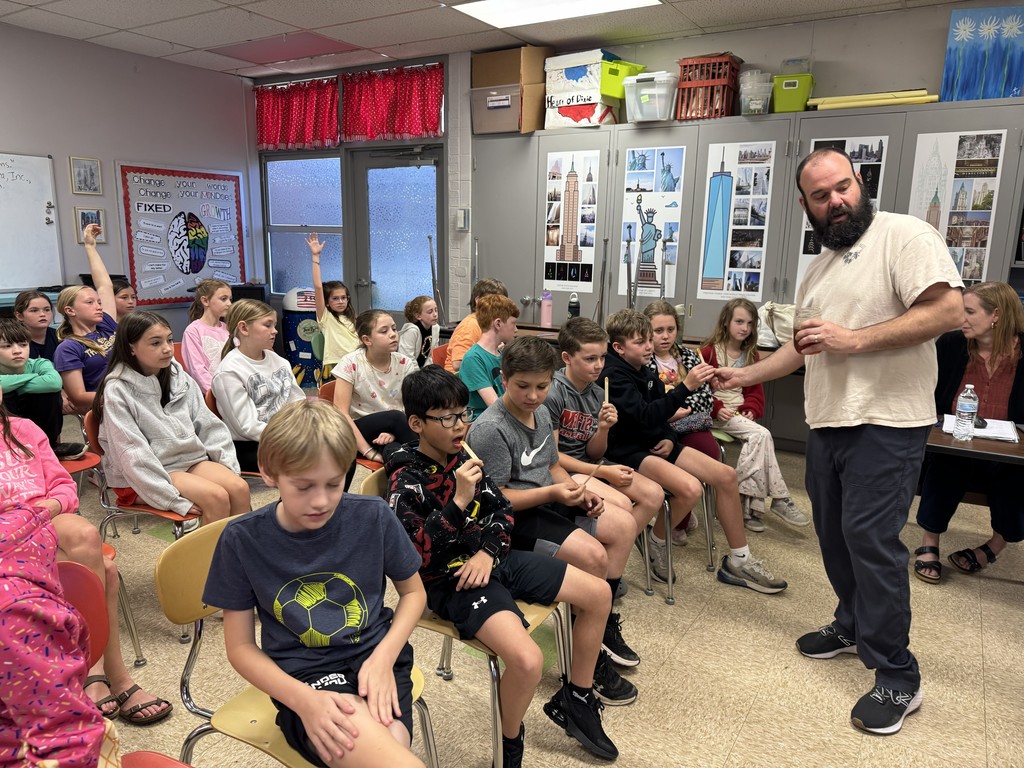 A visiting beekeeper looks on while 4th grade RLC students try a bite of honey on a craft stick. Students are raising their hands to volunteer to taste.
