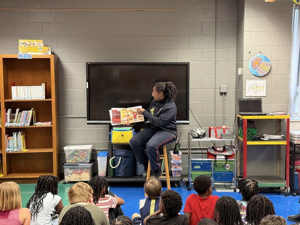 A firefighter sits on a stool and reads a book to a group of children inside a library. The students sit on the floor in front of the firefighter.