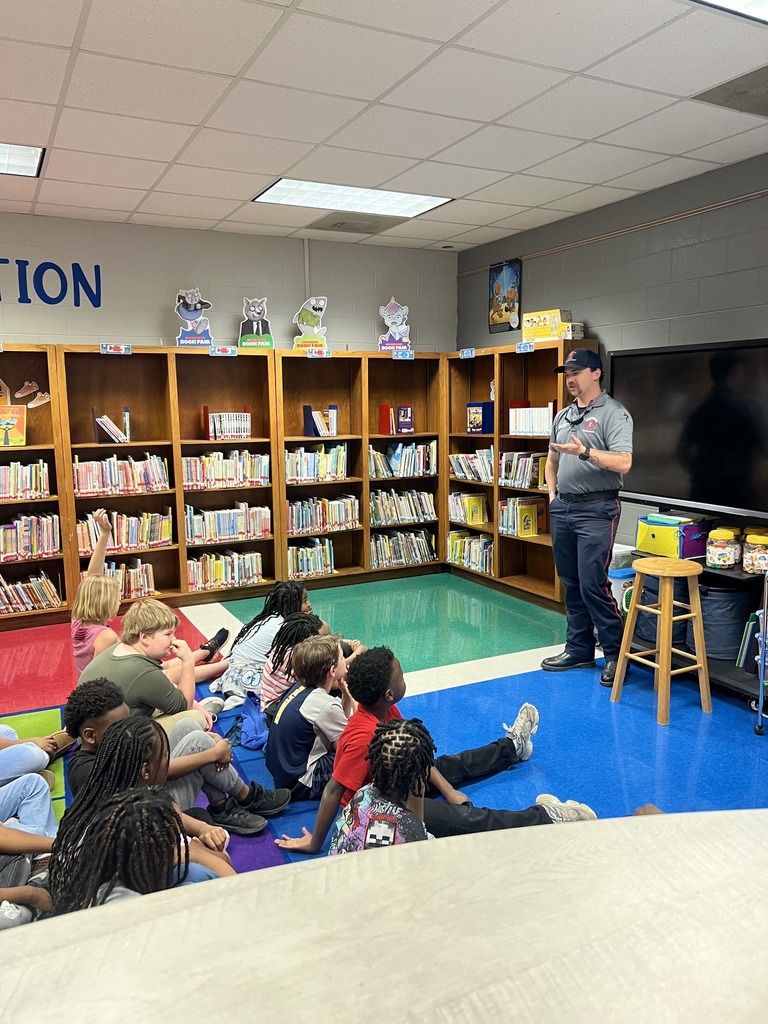 A firefighter stands in a school library and speaks to students as they sit on the floor.
