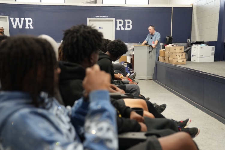 A man stands at a podium in the CCHS field house and speaks to football players  