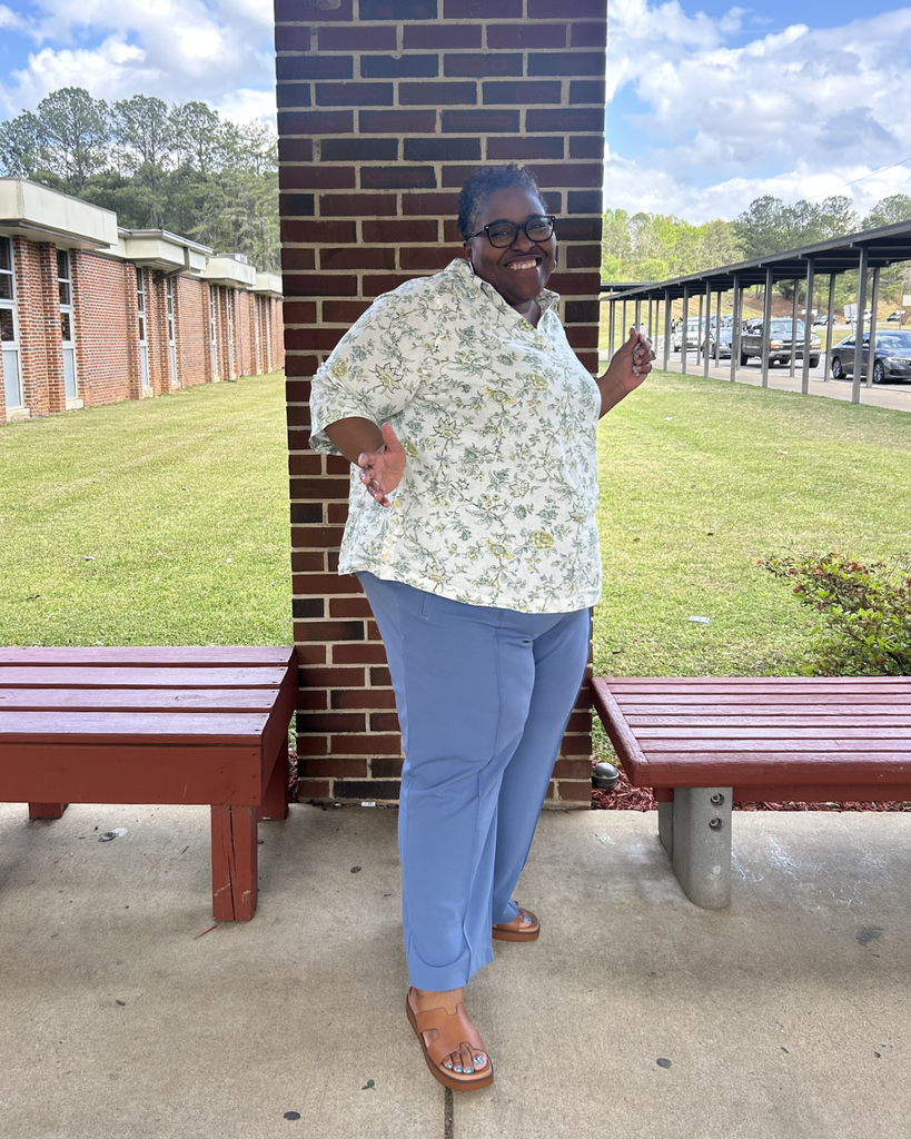 Krista Collins poses with a big smile outside Minor Middle School. She is leaning against a brick pillar between two benches, wearing a floral shirt and blue pants.