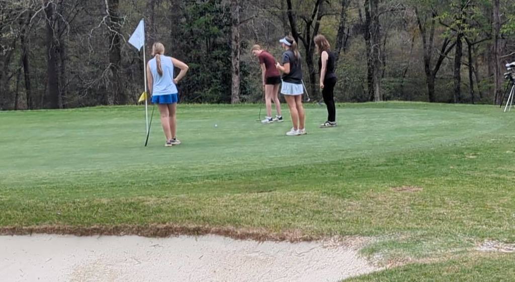Kennedy McAnnally gets ready to hit a golf ball with a golf club while competing at an outdoor course. Other golfers stand near her.