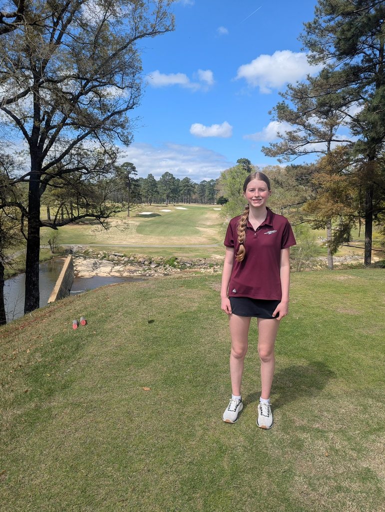 Kennedy McAnnally smiles for a photo while standing on a golf course. She wears a Gardendale High School golf polo.