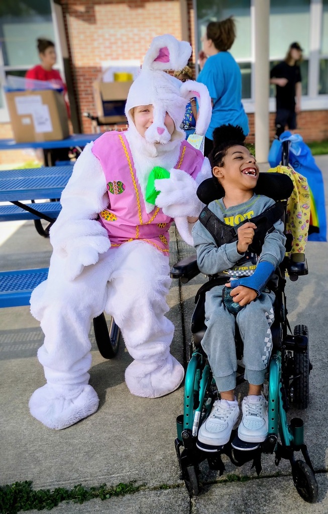 A person in a bunny costume poses next to a child outside. The child smiles and sits in a wheelchair.