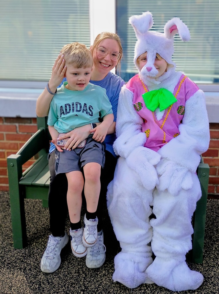 A person in a bunny costume sits on a bench outdoors next to an adult and child. All of them smile.