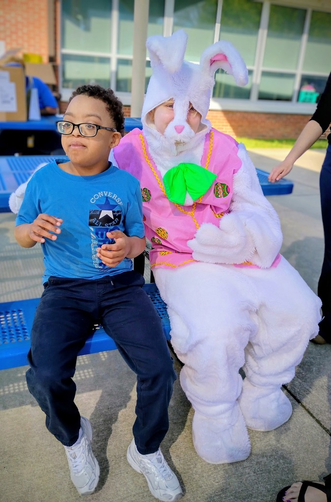 A person in a bunny costume sits outside at a picnic table and poses for a photo with a student. 