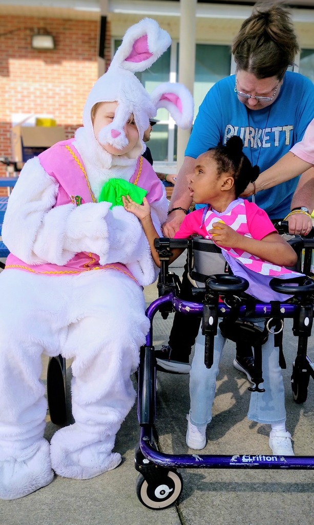 A person in a bunny costume sits next to a child at a picnic table. The child wears a pink shirt and jeans and uses a mobility aid.