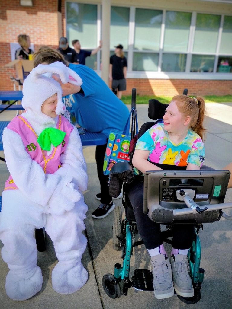 A person in a bunny costume sits outside and smiles at a child. The child wears a t-shirt with peeps on them and sits in a wheelchair.