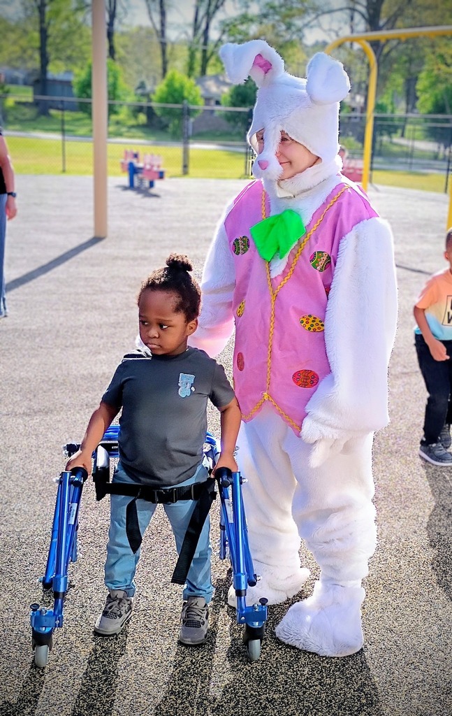 A person in a bunny costume stands with a child outside at a playground. The child stands and holds onto a walker.