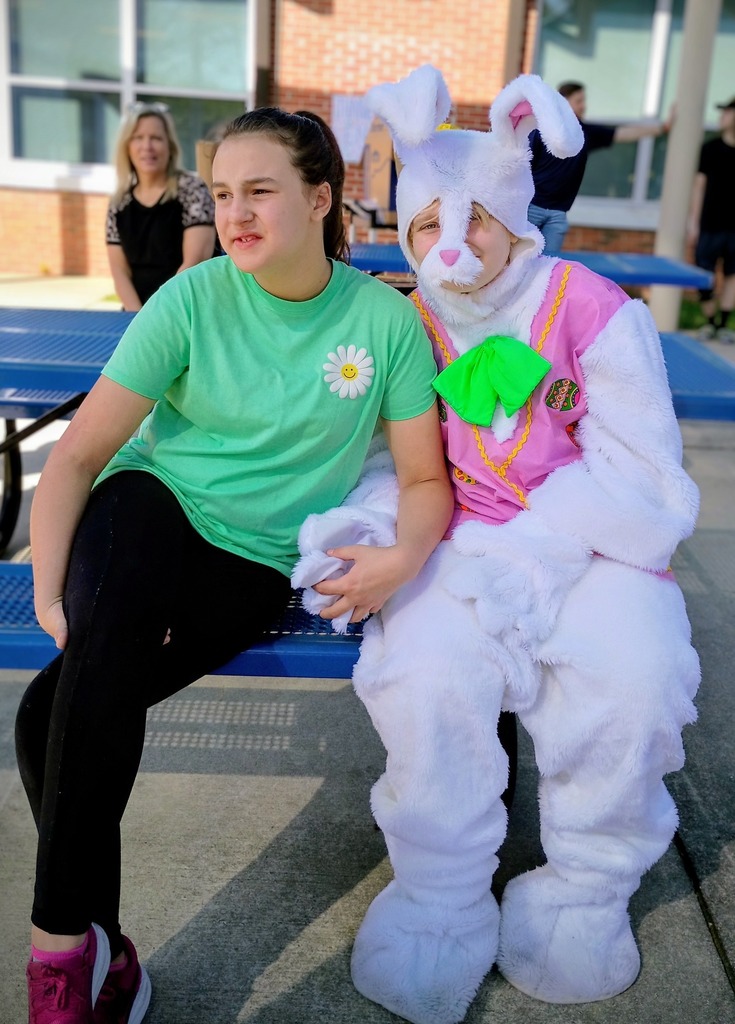 A person in a bunny costume sits at a picnic table and poses with a student. The student wears a green shirt and black pants and smiles.