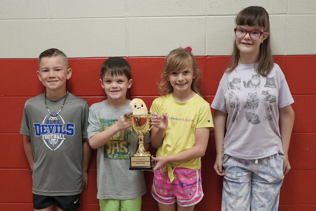 Four students stand together for a photo inside a school hallway. Two of the children hold up a trophy with a stuffed animal bean inside of it.