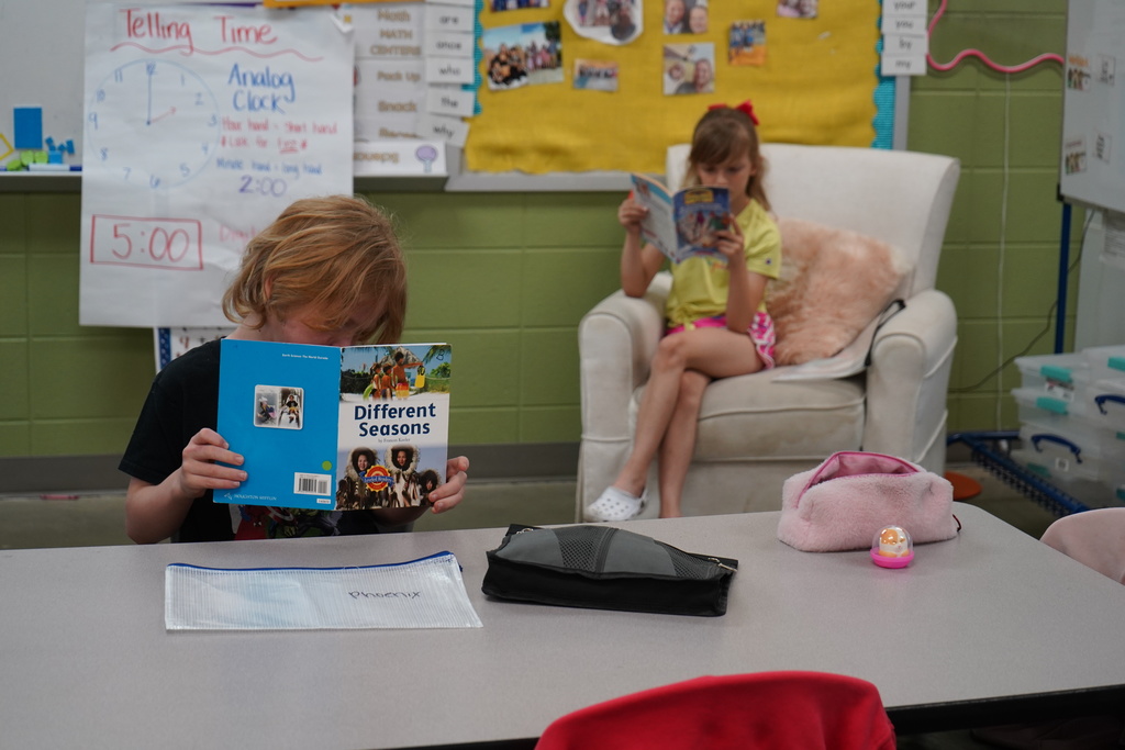 Two students read books inside a colorful classroom. One of the students sits at a table and the other sits in a chair.