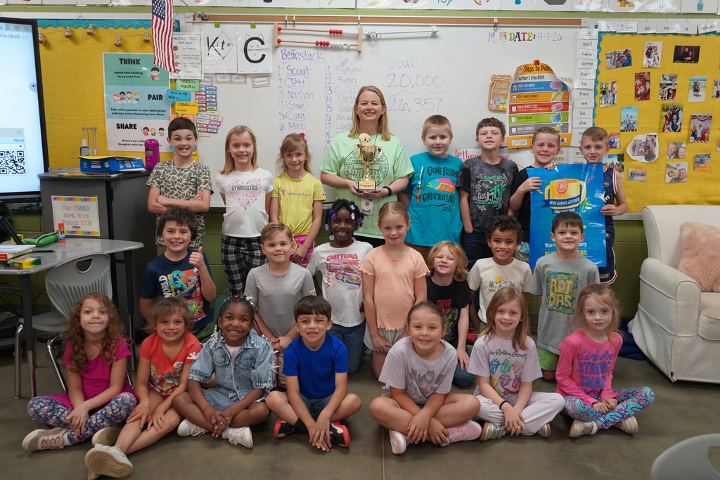 Students and a teacher stand and sit inside a classroom and pose for a photo. The children and adult all smile. Two of the children hold up a banner with a graphic of a trophy on it.