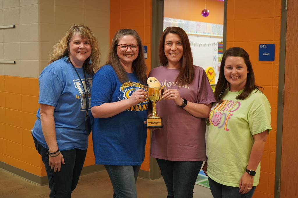 Four teachers stand together for a photo inside a school hallway. All of the teachers smile. Two of the teachers hold up a trophy with a toy bean inside.