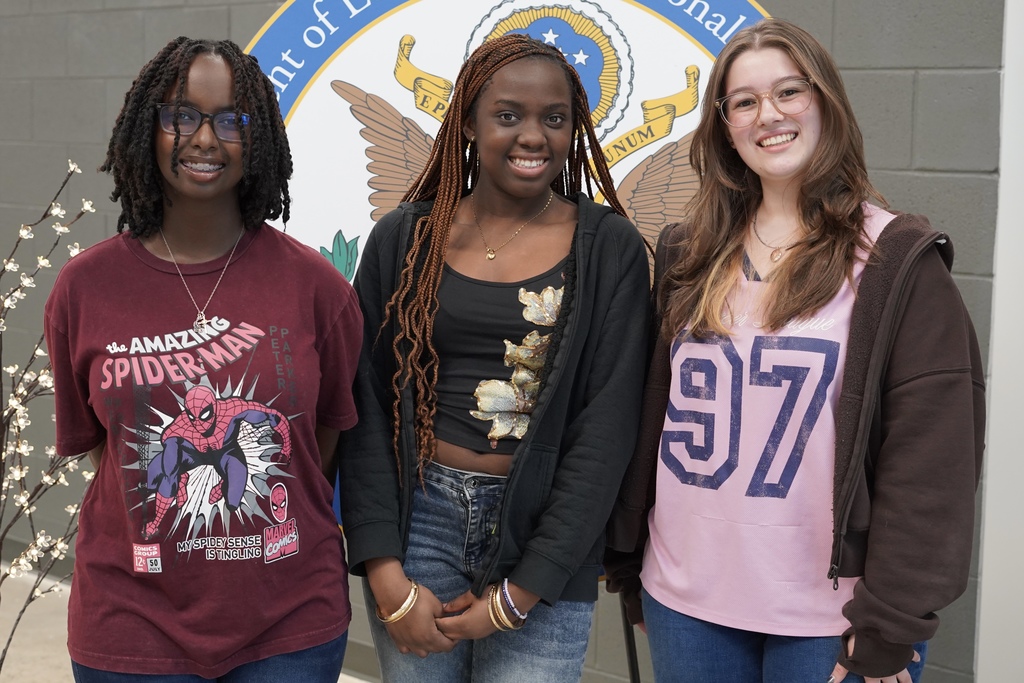 Three JCIB students stand together for a photo and smile. They stand inside the main entrance of the school and pose in front of a large sign with the school's seal on it.