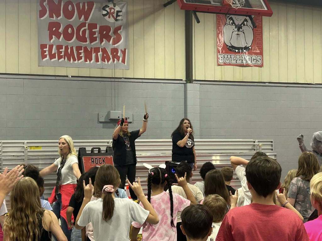 Two women stand in the Snow Rogers Elementary School gym and address a group of students. One woman holds drum sticks and the other talks into a microphone.