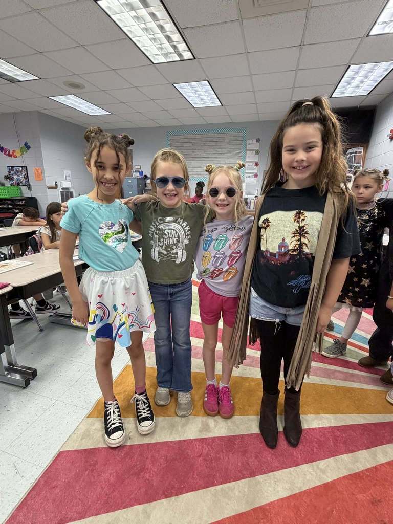 A group of students stand together for a photo in a classroom and smile. They wear rock and roll themed outfits.