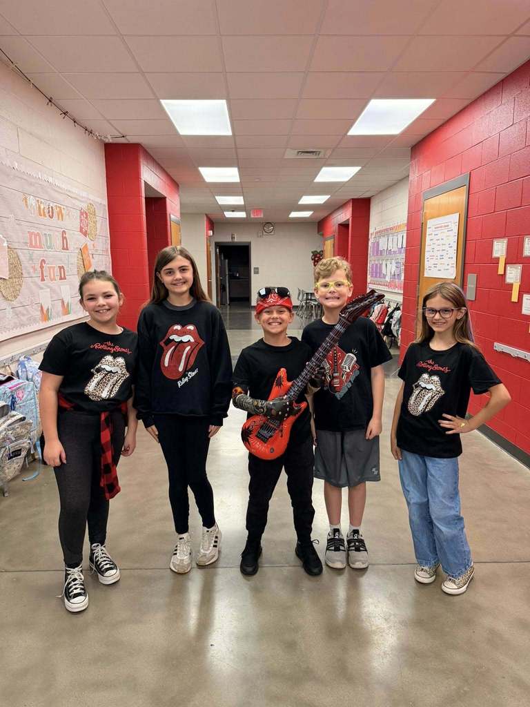 A group of students stand in a school hallway and pose for a photo. They wear rock and roll themed outfits and one student holds a guitar.