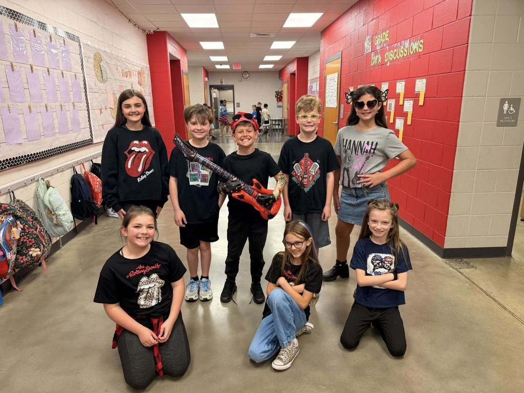 A group of students stand in a school hallway and pose for a photo. They wear rock and roll themed outfits and hold props.