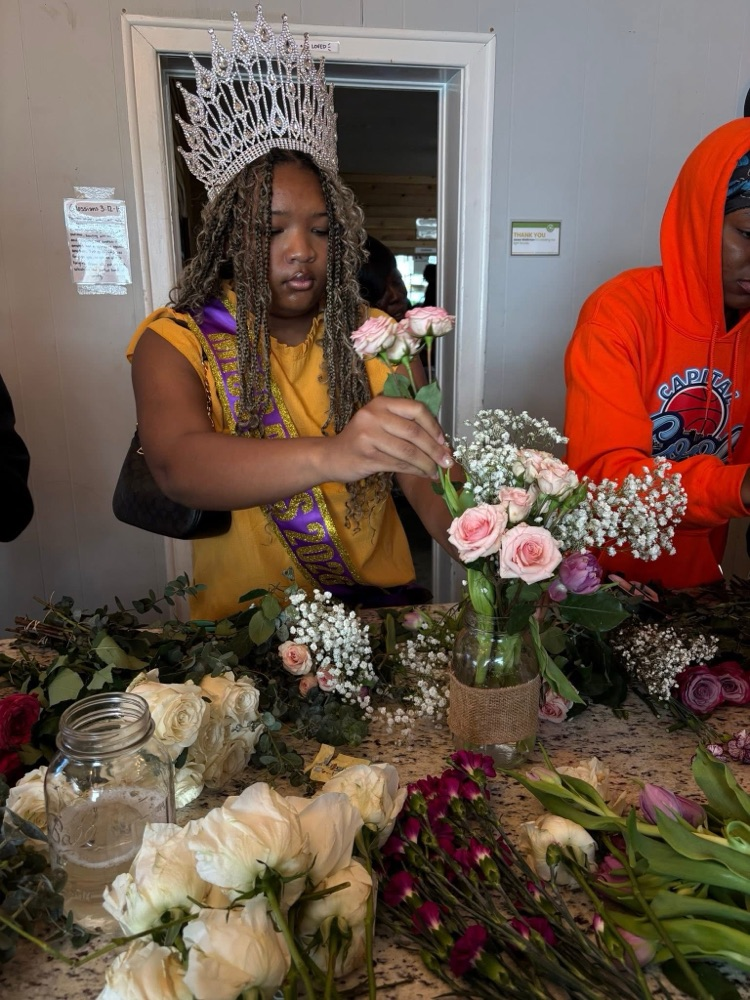A student wearing a crown and sash puts flowers into a vase. The student stands at a table covered with flowers inside a room  
