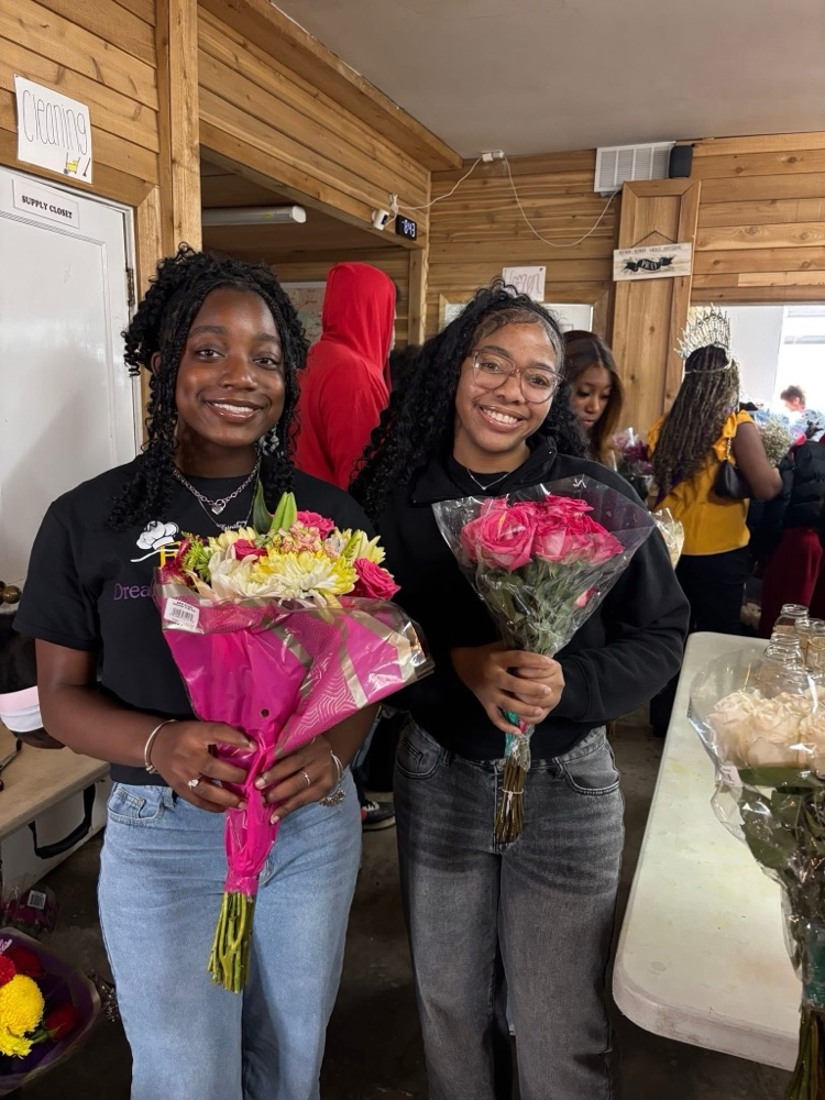 Two students smile while holding bouquets of flowers and standing inside a room  