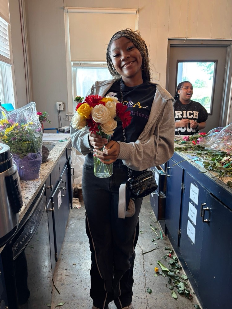 A student stands in a room and poses for a photo while smiling and holding a vase of flowers  