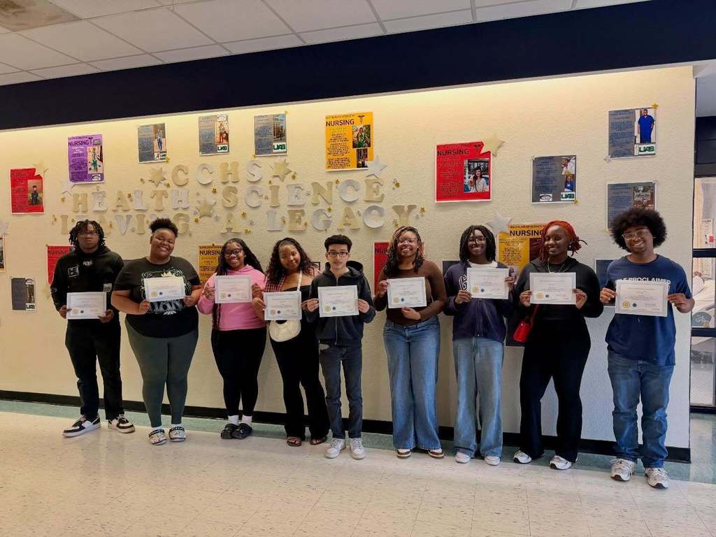 Students stand in a hallway of Clay-Chalkville High School and smile for a photo. They hold up certificates and stand in front of a wall that has letters spelling "CCHS Health Science Leaving a Legacy" on it, as well as photos of students and a description of their career path.