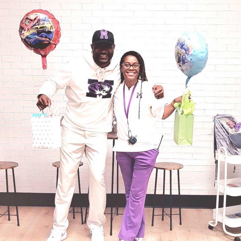 A smiling man and woman, identified as Mr. Phillips and Mrs. Steward, stand side-by-side in front of a white brick wall celebrating Assistant Principals Week. They are both dressed in school colors, wearing white tops and purple accents. Mr. Phillips wears a white hoodie with a purple school logo, a black baseball cap, and white pants, while Mrs. Steward wears a white polo and purple trousers. They are each holding a gift bag and a balloon—one red with race cars and one blue with characters—as they pose together with their arms around each other.