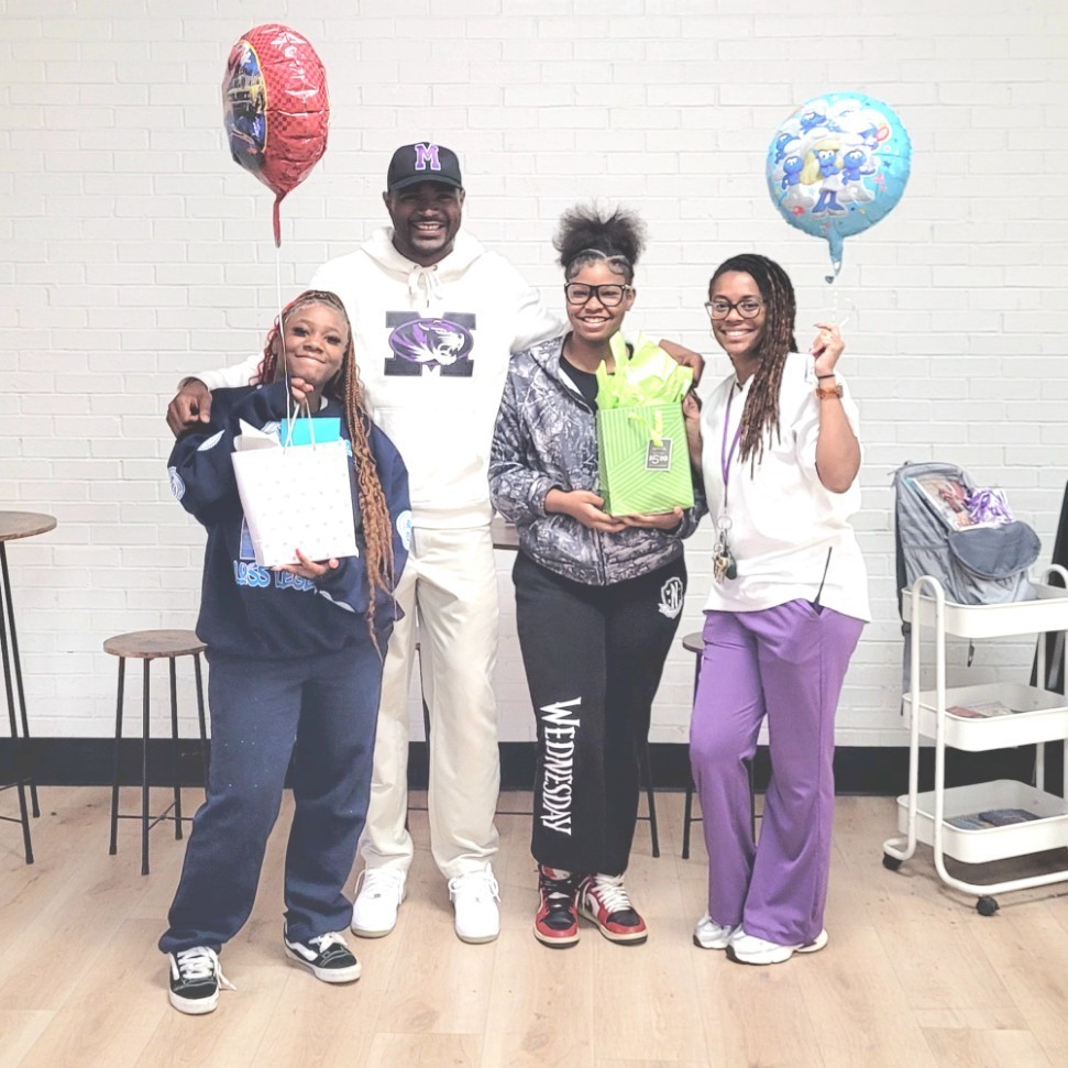 A group photo featuring Assistant Principals Mr. Phillips and Mrs. Steward smiling with two middle school students. They are standing against a white brick wall with a purple stripe at the top. Mr. Phillips stands in the center-back wearing a white school hoodie and a black "M" cap, with his arms around the students. To his left, a student holds a white gift bag and a red Hot Wheels balloon. To his right, another student holds a lime green gift bag. Mrs. Steward stands on the far right in a white polo and purple trousers, holding a blue Smurfs balloon. The group looks happy and cohesive, celebrating Assistant Principals Week.